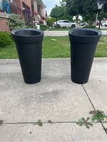 Photo shows two tall black tapered plastic outdoor planters standing on a concrete driveway.