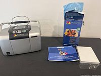 Photo showing the Epson PictureMate personal photo printer, user manual, extra ink cartridge box, and stacks of photo paper sheets.