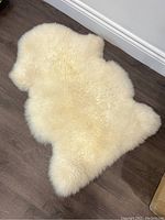 Cream colored sheepskin rug laid flat on dark wood floor, showing texture and shape.
