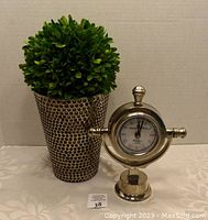 Front view of artificial green plant in hammered metallic pot with circular indentations and silver-tone decorative clock with round face and Roman numerals.