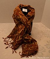 Fashion scarf displayed wrapped around a black mannequin bust showing rich brown and rust tones with a heavy gauze texture and knotted tassel fringe ends.