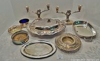 Photo showing the full lot of silver plated items arranged on a table, highlighting the candlesticks, chafing dish, trays, bowls, and wine bottle stands.