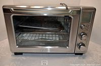 Front view of the Oster toaster oven showing stainless steel body, glass door, and knobs on right side.