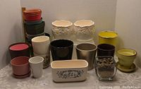 Photo shows a variety of plant pots in different sizes, colors, and materials arranged on a table. Includes stacked plastic pots, ceramic pots with floral designs, and a glass container with rocks.