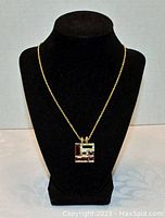 Front view of the pendant necklace displayed on a black bust stand, showing gold-tone chain and square pendant with embedded crystals.