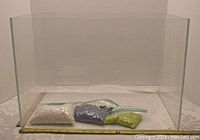 Clear glass terrarium with three bags of colored gravel inside and placed in front of it. Measuring tape in front shows approximate dimensions.