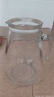 Clear glass pitcher with handle and pouring spout shown from an angle.