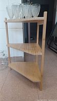 Front angle view of small three-tier wooden corner shelf with glass cups on top shelf.