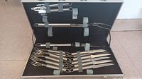 Photo of the stainless steel steak knives, forks, cooking knife, meat fork, and can opener secured inside a black travel case with straps.