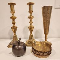 Front view showing two brass candle holders, brass chalice/vase, dark metal apple bell, and eight brass ashtrays/coasters stacked