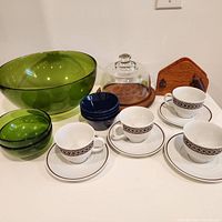 Full view of lot showing large green glass salad bowl, two small matching green bowls, five blue pinch bowls stacked, four white porcelain cups with black and brown geometric design and matching saucers, wooden napkin holder and glass cake dome on wood base.
