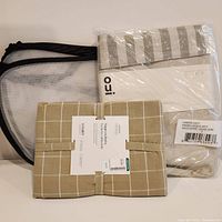 Three items on a white surface: organic cotton tablecloth with beige and white grid pattern, Paige Kessley 2021 hamper in packaging with gold ochre stripes, and mesh hamper with black frame