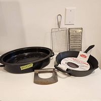 Cast iron skillet with label inside next to large oval roasting pan, vintage meat cleaver and two vintage kitchen tools placed on white surface