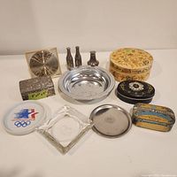Photo displays all items in the lot arranged on a white surface including salt shaker, box with blue velvet lining, coaster set in paper mache box, vintage tin bowl, glass ashtrays, small tins and a collectible Olympic plate.