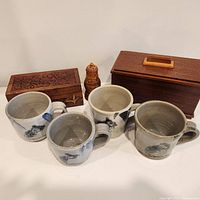 Front view of four pottery mugs with bird designs along with two wooden tea boxes and a carved wooden salt shaker representing entire lot