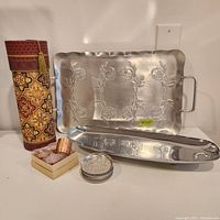 Overview photo showing all items: cardboard wine tube, embossed aluminum serving dish, long aluminum tray, copper napkin holders in box, and stack of ashtrays/coasters