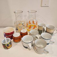 Vintage glass juice jugs with fruit patterns, orange ceramic salt and pepper shakers, various Dansk mugs, jam/sugar pot with spoon, and red pinch pots arranged on a white surface.
