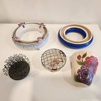 Top view of two ceramic flower rings, one white with pink roses and one blue and white, two metal flower frogs, and a floral wall vase.