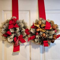 Two artificial snowy pine wreaths hanging on red fabric ribbons over a white door, showing natural nuts, acorns, seed pods, and red bows.