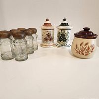 Front view of 3 ceramic lidded spice pots with decorative botanical patterns beside 6 glass spice containers with wood lids.