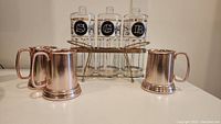 Front view of three Roman-themed glass liquor decanters with black circular labels 'S', 'G', 'R' sitting in a silver metal caddy with three light metal steins arranged around the caddy.
