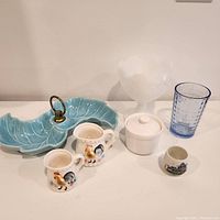 Front view of all vintage kitchenware items including serving dish, measuring cups, compote glass, lidded jar, saki/toothpick holder, blue glass, and pinch pot.