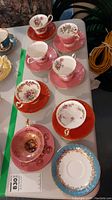 Seven Aynsley tea cups with matching saucers, displayed on a table, showing variety of floral designs and colors such as red, pink, and blue backgrounds.