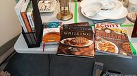 Photo showing six cookbooks on a table with a black mesh metal book holder beside white ceramic dishes.