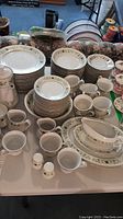 Full view of the set arranged on a tabletop showing stacks of plates, bowls, cups, jugs, covered sugar bowls, gravy boat, salt and pepper shakers, and coffee pot all decorated with green floral vine border pattern on white porcelain.