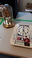 Photo showing the anniversary clock with gold-tone base and plastic dome alongside the illustrated wall clock resting on a table.