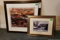 Shows two framed prints side by side on carpeted floor, one fall scene with dark frame and one winter scene with gold frame