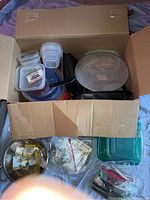 Open cardboard box filled with various plastic Tupperware containers with snap lids, black cooking utensils, and a mesh strainer visible inside and surrounding the box.