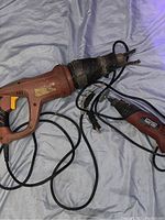 Two electric power tools laid out on grey fabric: an electric saw and a sander/grinder with cords