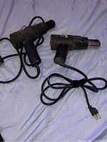 Two black electric heat guns with cords on a blue fabric surface, showing overall size and design.