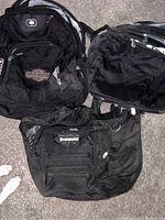 Three black backpacks laid on carpet. One Harley Davidson with logo patch, one Brunswick, one Fisher Price backpack showing front mesh pocket.
