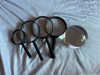 Five magnifying glasses shown on white cloth, includes one glass dome ball and four handheld magnifiers with black handles.