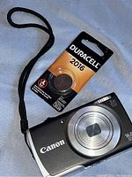 Canon digital camera with wrist strap and Duracell 2016 lithium battery still in original packaging displayed on a light surface