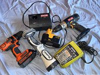 Photo showing two Ryobi cordless drills, Makita IX7 flashlight, two Ryobi battery chargers and two rechargeable batteries displayed on a cloth background.
