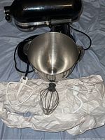 KitchenAid Artisan black stand mixer head and large stainless steel mixing bowl with three attachments (flat beater, wire whip, dough hook) laid out on white paper.