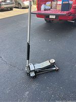 Metal floor jack with long handle and rubber grip placed on asphalt surface by the open back of a red vehicle.