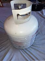 Side angle view of the white propane tank showing body and protective collar around the valve with some surface wear.