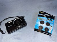 Samsung black digital camera with 5x zoom lens, LCD screen, and a wrist strap. Pack of four CR2032 lithium button cell batteries in blue/white packaging shown beside camera.