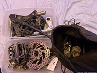 Photo of various bicycle parts in a clear plastic bin and black fabric bag including pedals, metal kickstands, disc brake rotors, and assorted metal parts.
