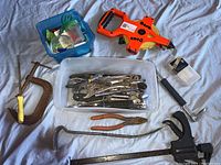 Assorted hand tools including large orange tape measure, socket wrench set in plastic bin, C-clamp, pliers, screwdrivers, and metal ruler on gray cloth