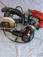 Overview of three tools: Ryobi circular saw in black and silver, two red Craftsman corded electric drills placed on a blue fabric surface.