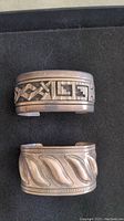 Top view of both cuff bracelets on black background showing geometric inlay and wave repoussé designs