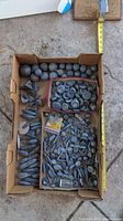 Overhead image of large assortment of various shaped lead fishing weights laid in cardboard box. Includes balls, pyramids, teardrop shapes, discs and cylinders.