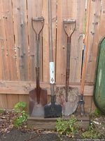 Four garden tools standing upright against a wooden fence: spade, edger or ice scraper, shovel, and cultivator with wooden handles and metal heads showing signs of use.