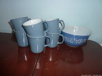 Six pale blue mugs with white interiors stacked and arranged next to a blue mixing bowl with white floral pattern.