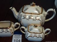 Three-piece Sadler Cube tea set comprising teapot, sugar bowl, and creamer shown adjacent with auction tag. White to off-white with gold trim and decorative patterns.